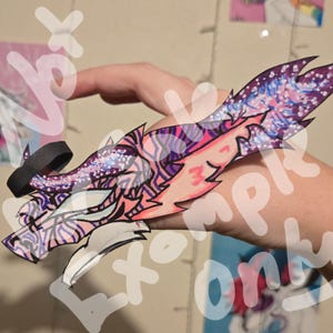 May include: A hand-drawn paper craft of a purple, pink, and blue dragon with glitter accents. The dragon has a black ring around its neck and is holding its head up.