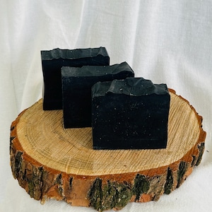 May include: Three rectangular bars of solid black soap with textured, uneven tops. The soap bars are arranged in a staggered formation, showcasing a deep, matte black colour throughout each piece, providing a stark contrast to the natural wood base.