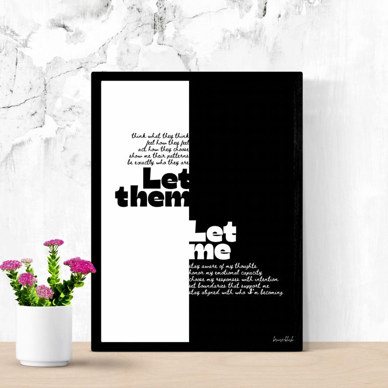 May include: A framed art print with a black and white design. The artwork features the words "Let them" and "Let me" in bold, white font. Additional text is included in a smaller, cursive font. A small white pot with pink flowers sits in front.