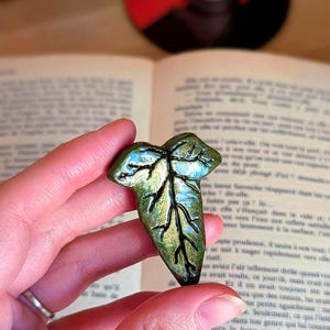 Peut inclure: Une broche en forme de feuille avec une finition brillante, présentant un dégradé de teintes or métallique et vert profond. La surface est détaillée avec des nervures noires en relief créant un aspect organique et texturé sur la feuille.