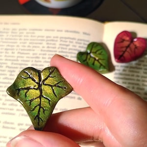 Peut inclure: Trois ornements en forme de feuille en cœur avec une finition brillante. L'un est tenu au premier plan, présentant un dégradé de vert et d'or avec des nervures sombres gravées. Deux autres ornements, un vert et un rouge, sont posés sur un livre.