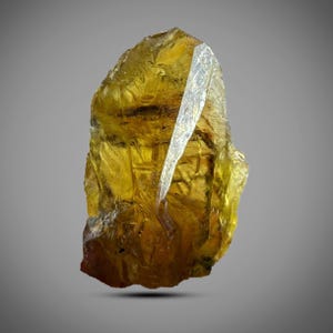 Könnte beinhalten: Ein durchscheinender, bernsteinfarbener Kristall mit einer gezackten, unregelmäßigen Form. Ein klarer, kristalliner Splitter ragt aus der Spitze heraus und erzeugt einen auffälligen Kontrast. Der Edelstein ist vor einem neutralgrauen Hintergrund platziert.