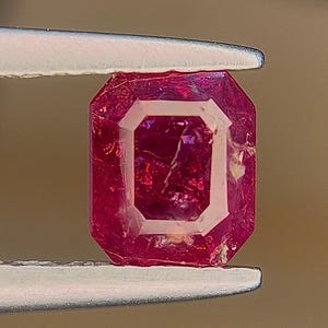 Può includere: Un rubino sfaccettato, rettangolare, tenuto da una pinzetta. Il rubino è di colore rosso intenso con un design interno rettangolare più chiaro. La pinzetta è argentata e lo sfondo è marrone sfocato.