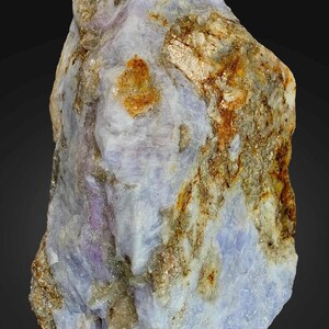May include: A natural gemstone with a mix of pale blue, lavender, and brown hues. The rock's surface has a textured appearance, with areas of metallic sheen and patches of rust-coloured minerals. The overall shape is irregular, showcasing its raw, unpolished state.