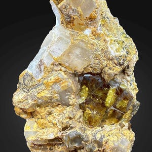 Könnte beinhalten: Ein rohes, ungeschnittenes Mineralexemplar mit einer Mischung aus Farben. Das Stück weist durchscheinende, hellfarbige Kristallformationen neben Bereichen in Goldgelb- und Brauntönen auf. Die Gesamttextur wirkt rau und natürlich, vor dunklem Hintergrund.