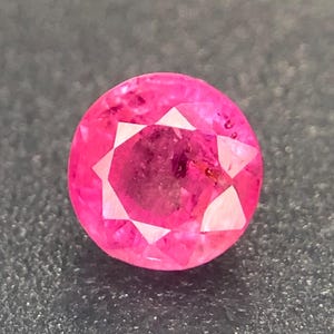 May include: A round, faceted gemstone with a vibrant pink hue. The gem features a central dark area surrounded by a series of white, triangular facets, creating a starburst effect. The gemstone is set against a dark, textured background.
