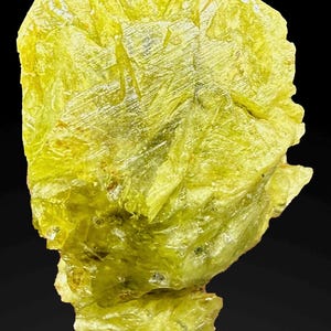 Könnte beinhalten: Nahaufnahme eines leuchtend gelben Schwefelkristallclusters vor schwarzem Hintergrund. Das Mineral hat eine raue, geschichtete Textur mit kristallinem Aussehen. Die Farbe ist ein helles, fast neonfarbenes Gelb.