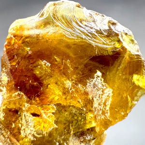 Könnte beinhalten: Nahaufnahme eines durchscheinenden, bernsteinfarbenen Edelsteins. Der Stein hat eine raue, unregelmäßige Form mit glänzender Oberfläche. Licht reflektiert sich auf der Oberfläche und erzeugt Glanzlichter und Schatten. Der Edelstein scheint ein natürliches Mineralexemplar zu sein.