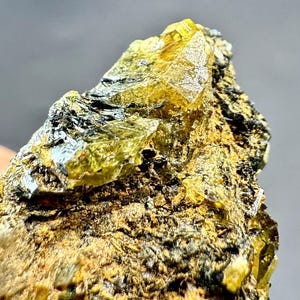 Könnte beinhalten: Nahaufnahme einer rauen, natürlichen Mineralprobe. Der Stein weist eine Mischung aus Texturen und Farben auf, mit markanten gelb-grünen Kristallformationen. Die Basis ist eine Mischung aus Braun und Schwarz, was einen auffälligen Kontrast erzeugt.