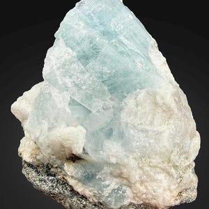 Puede incluir: Un espécimen de cristal de aguamarina en bruto. La gema presenta una estructura cristalina azul pálido con matriz blanca y gris. La aguamarina tiene una apariencia translúcida y está sobre un fondo oscuro.