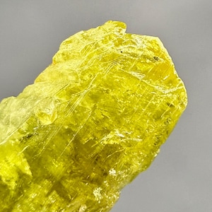 Könnte beinhalten: Nahaufnahme eines leuchtend gelben Schwefelkristalls. Die Kristallstruktur hat eine raue, strukturierte Oberfläche mit sichtbaren Streifen. Das Mineral ist vor einem grauen Hintergrund platziert, der seine lebendige Farbe und natürliche Form hervorhebt.