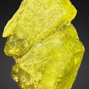 Könnte beinhalten: Nahaufnahme einer leuchtend gelben Schwefelkristallformation. Die kristalline Struktur hat eine raue, strukturierte Oberfläche und ein lebendiges, fast durchscheinendes Aussehen. Das Mineral ist vor einem dunklen Hintergrund platziert.