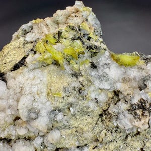 Könnte beinhalten: Nahaufnahme einer Mineralprobe mit leuchtend gelben Kristallen. Die Kristalle heben sich von einem Hintergrund aus weißen, grauen und schwarzen Gesteinsformationen ab, was einen auffälligen Kontrast erzeugt. Die Textur ist rau und uneben, was die natürliche Schönheit des Minerals hervorhebt.