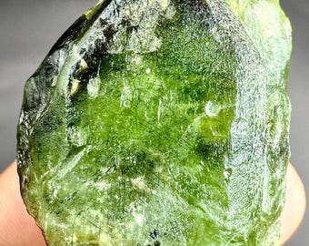 Puede incluir: Una gema de peridoto en bruto, sin cortar, de un vibrante color verde. El cristal tiene una superficie rugosa y texturizada con inclusiones oscuras y una parte superior puntiaguda. La piedra se sostiene en una mano, mostrando su forma y color naturales.