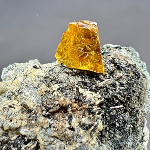 Könnte beinhalten: Nahaufnahme einer rauen, goldgelben Mineralprobe. Der durchscheinende, bernsteinfarbene Kristall sitzt auf einer dunklen, strukturierten Felsformation. Das Mineral hat eine zerbrochene, unregelmäßige Form, und der Fels hat ein gesprenkeltes Aussehen.