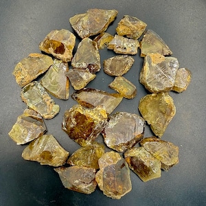 Könnte beinhalten: Eine Sammlung roher, ungeschliffener Rauchquarzkristalle. Die Kristalle variieren in Größe und Form und weisen eine Reihe von durchscheinenden bis undurchsichtigen braunen und bernsteinfarbenen Farbtönen auf. Die raue, natürliche Textur der Steine ist sichtbar.