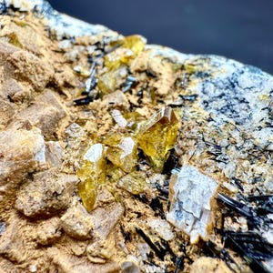 Könnte beinhalten: Nahaufnahme einer Mineralprobe mit goldgelben Kristallen und schwarzen nadelförmigen Formationen, eingebettet in eine beige und weiße Matrix. Die Kristalle haben ein durchscheinendes Aussehen mit scharfen Kanten.