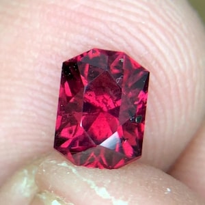 Puede incluir: Una gema facetada de color rojo intenso con forma rectangular. La gema tiene múltiples facetas que reflejan la luz, creando un efecto brillante. La piedra se sostiene entre los dedos, mostrando su color y claridad.
