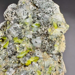 Könnte beinhalten: Nahaufnahme einer Mineralprobe mit einer Matrix aus grauem und schwarz gesprenkeltem Gestein. Eingebettet sind durchscheinende, rechteckige Kristalle und leuchtend limettengrüne, längliche Kristalle. Die Zusammensetzung ist natürlich und rau.