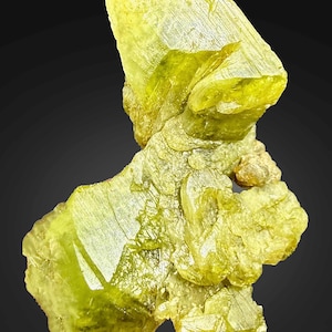 Könnte beinhalten: Nahaufnahme eines leuchtend limettengrünen Mineralexemplars. Die kristalline Struktur hat ein geschichtetes Aussehen mit scharfen Kanten und einer rauen Textur. Das Mineral ist vor einem dunklen Hintergrund platziert, der seine Farbe und Form hervorhebt. Dies ist ein natürlicher Edelstein.