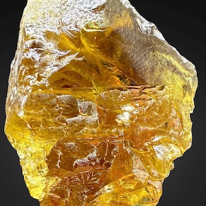 Könnte beinhalten: Ein großer, grob geschliffener Edelstein in goldgelber Farbe. Die Oberfläche ist strukturiert mit sichtbaren Brüchen und einem kristallinen Aussehen. Der Stein ist vor einem dunklen Hintergrund platziert.