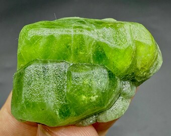 Puede incluir: Una vibrante piedra preciosa de peridoto verde translúcido con una estructura cristalina facetada. El mineral en bruto y sin cortar muestra una forma natural e irregular y un color verde lima brillante, que refleja la luz. La piedra preciosa se sostiene en una mano.