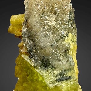 Könnte beinhalten: Nahaufnahme einer Mineralprobe mit kristalliner Struktur. Das Mineral weist einen Farbverlauf auf, der von einem leuchtenden Gelb an der Basis zu einer klaren, kristallinen Spitze übergeht. Dunklere Einschlüsse sind sichtbar.