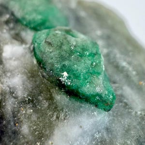 Puede incluir: Primer plano de una gema de esmeralda en bruto, sin cortar, con un tono verde vibrante. La esmeralda se encuentra sobre un fondo de roca gris y blanca, mostrando su textura natural y estructura cristalina.