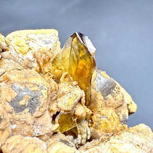 Könnte beinhalten: Nahaufnahme eines Citrin-Kristallclusters. Der Kristall hat eine durchscheinende gelbbraune Farbe mit einer scharfen, spitzen Spitze. Er ist vor einem Hintergrund aus rauem, beigefarbenem Gestein mit grauen und gelben Flecken angeordnet.