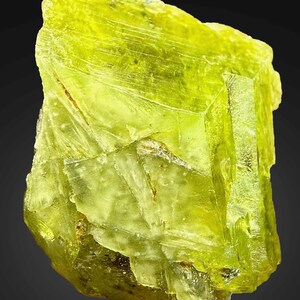 Könnte beinhalten: Ein lebendiges, durchscheinendes, gelb-grünes Mineralexemplar mit kristalliner Struktur. Die rauen Kanten und Facetten fangen das Licht ein und zeigen die natürliche Schönheit des Steins. Die Farbe des Minerals ist ein heller, auffälliger Farbton.