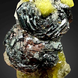 Könnte beinhalten: Nahaufnahme einer Mineralprobe mit dunklen, metallisch aussehenden Kristallen in einer geschichteten Struktur. Die Kristalle werden durch leuchtend gelb-grüne Formationen akzentuiert, was einen auffälligen Kontrast erzeugt. Das Mineral sitzt auf einer hellen Basis.