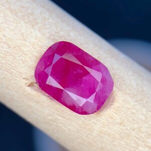 May include: A faceted, rectangular ruby gemstone with a vibrant, deep pink hue. The gem is held against a light-coloured wooden surface, showcasing its polished facets and internal brilliance. The gemstone's shape is a modified cushion cut.