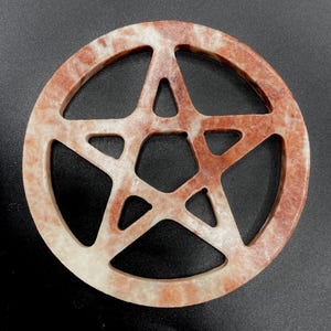 Könnte beinhalten: Ein kreisförmiges, durchbrochenes Pentagramm in Rot- und Cremetönen. Der fünfzackige Stern ist aus einem steinartigen Material mit einem gesprenkelten Muster geschnitzt. Das Objekt befindet sich vor einem dunklen Hintergrund.