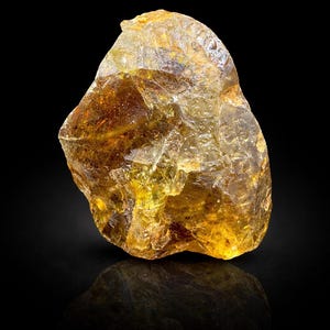 Könnte beinhalten: Ein roher, ungeschliffener Edelstein mit goldgelber Farbe. Das durchscheinende Mineral hat eine kristalline Struktur und ist vor einem schwarzen Hintergrund platziert, mit einer Spiegelreflexion darunter. Die Oberfläche des Steins ist uneben und strukturiert.