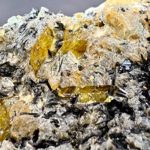Könnte beinhalten: Nahaufnahme eines Mineralexemplars mit durchscheinenden gelben Kristallen und schwarzen, nadelförmigen Einschlüssen. Das Mineral hat eine raue, strukturierte Oberfläche mit grauen und weißen Bereichen.