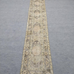 May include: A long, rectangular rug with an intricate, faded floral pattern in shades of beige, brown, and olive green. The rug's design features repeating medallions and borders, creating a vintage or antique aesthetic. The rug is suitable for a hallway or entryway.