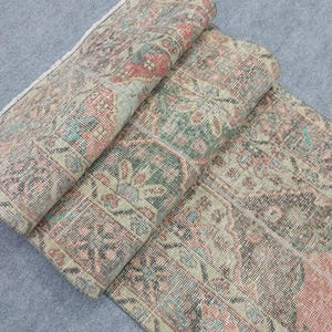 May include: A vintage-style rug with a distressed look. The rug displays a detailed floral pattern in faded red, green, and blue hues against a beige backdrop. The rug is partially rolled, showcasing its intricate design and texture.