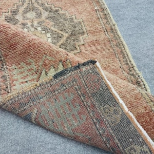 May include: A folded, vintage rug with a distressed, faded appearance. The rug features a geometric pattern in shades of peach, brown, grey, and black. The edges are frayed, and the rug appears to be made of woven material.