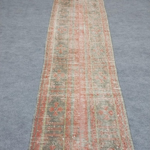 May include: A long, rectangular rug with a distressed, vintage appearance. The rug features a faded color palette of red, gray, and cream, with a repeating geometric pattern. The rug is likely made of woven material and is suitable for a hallway or entryway.