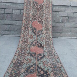 May include: A long, rectangular rug with an intricate, symmetrical pattern. The rug features a central design in shades of coral and black, framed by borders in muted greens and browns. The rug is displayed outdoors, leaning against a stone wall.