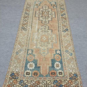 Puede incluir: Una alfombra rectangular con un medallón geométrico central en tonos melocotón, crema y azul apagado. El borde muestra un patrón floral repetitivo con líneas intrincadas y formas simétricas, exhibiendo una textura desgastada y descolorida.