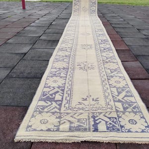 May include: A long, rectangular rug with a cream background and a repeating blue geometric pattern. The rug is displayed outdoors, showcasing its length and design. The edges have a fringed detail.