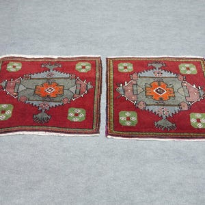 Può includere: Due tappeti quadrati abbinati con sfondo rosso e intricati motivi geometrici. Il design centrale presenta una forma a diamante con accenti arancioni e grigi, circondata da motivi floreali verdi e bianchi. I tappeti hanno un bordo con frange.