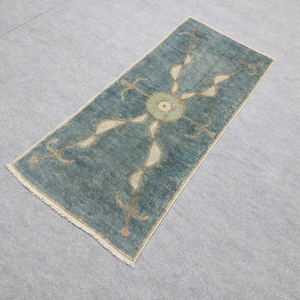 May include: A rectangular rug with a teal background and a central design featuring a circular element and flowing, ribbon-like shapes in beige and gold. The rug has a fringed edge.