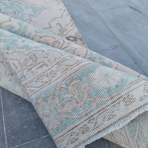 Puede incluir: Una alfombra enrollada de estilo vintage con aspecto desgastado. La alfombra presenta una base color crema con patrones florales descoloridos en tonos turquesa y marrón. Los bordes tienen flecos y la alfombra está parcialmente desenrollada.