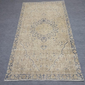 Puede incluir: Alfombra rectangular beige con un diseño central de medallón en azul marino y marrón claro. La alfombra presenta un intrincado patrón floral y un borde azul marino. La alfombra está colocada sobre una superficie gris.