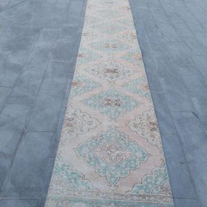 May include: A long, rectangular rug with a faded, vintage appearance. The rug features a central design of repeating teal and brown floral medallions on a light peach background. The border has a similar teal and brown pattern.