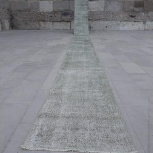 May include: A long, narrow, rectangular rug with a faded, distressed appearance. The rug is primarily a muted green color with lighter, faded areas, and a fringe at one end. The rug is placed on a gray stone surface, with a stone wall in the background.