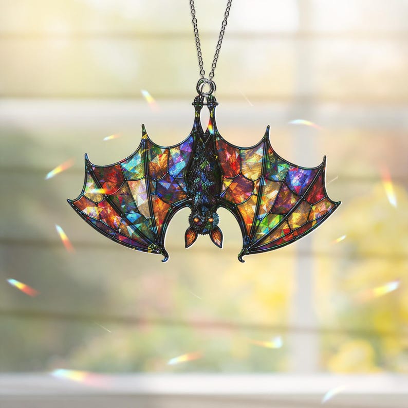 May include: A hanging ornament shaped like a bat with wings featuring a multi-colored stained glass pattern in shades of red, blue, green, and yellow. The bat has a dark body, orange eyes, and is suspended from a silver-toned metal chain.