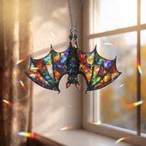 May include: A hanging ornament shaped like a bat with wings featuring a multi-colored stained glass pattern. The bat is dark with orange eyes and is suspended by a silver-toned metal chain. The wings display a mosaic of vibrant colors.
